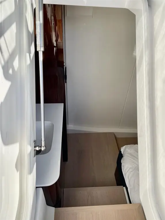 Interior view of a yacht, showing a staircase, a bedside, and a table under natural light. Wood finishes and white walls visible.